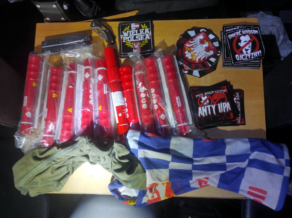 Na drewnianym blacie leżą różne przedmioty, w tym pakiety czerwonych rac, zapalniczka, plakaty z hasłami oraz kawałek odzieży.