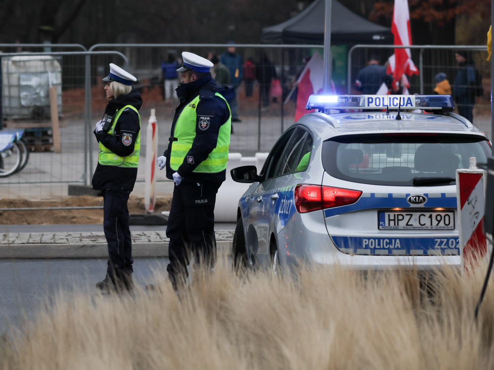 Dwóch policjantów w kamizelkach odblaskowych rozmawia obok radiowozu policyjnego na ulicy.