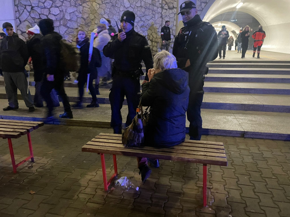 Kobieta siedząca na ławce rozmawia z dwoma policjantami w ruchliwym miejscu, wokół przechodnie.