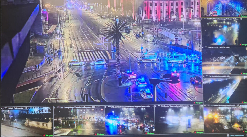 Policyjne radiowozy z włączonymi sygnałami świetlnymi blokują ruch na mokrej ulicy, w tle widoczne oświetlenie miejskie.