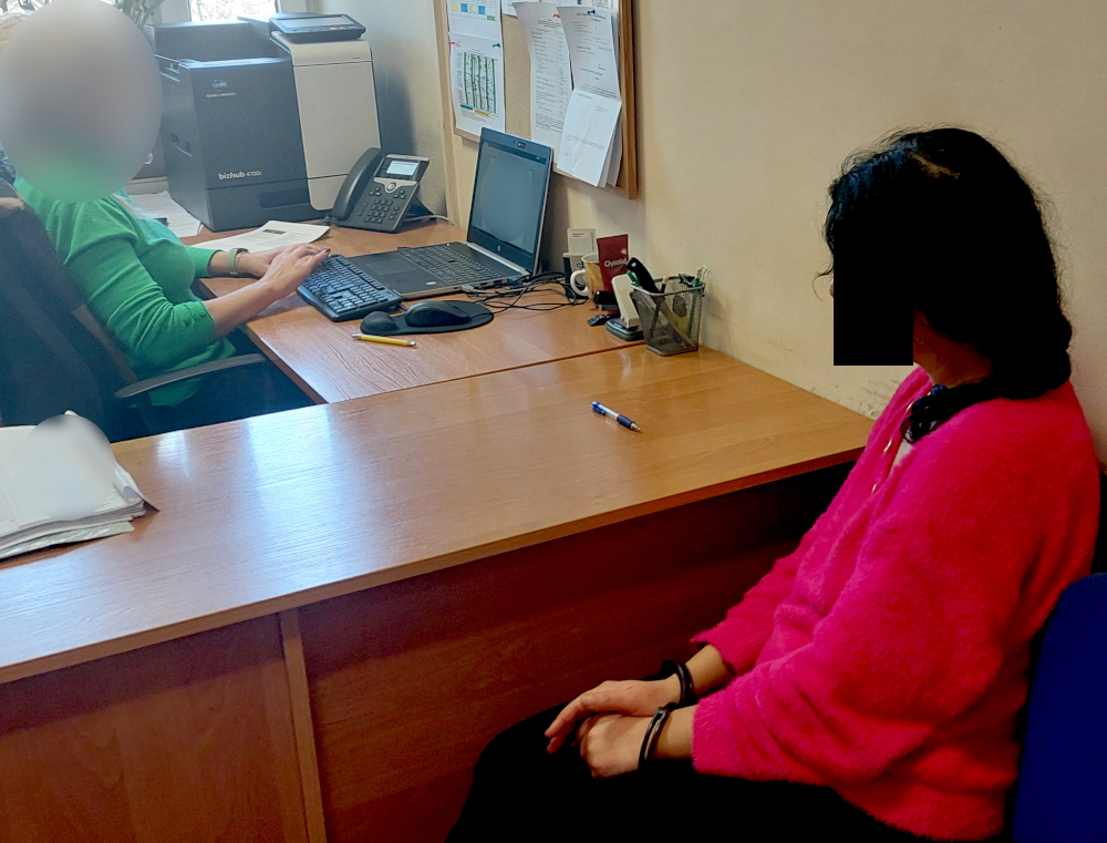 Kobieta w różowej bluzie siedzi na krześle, obserwując pracującą kobietę w zielonej bluzce przy biurku z komputerem.