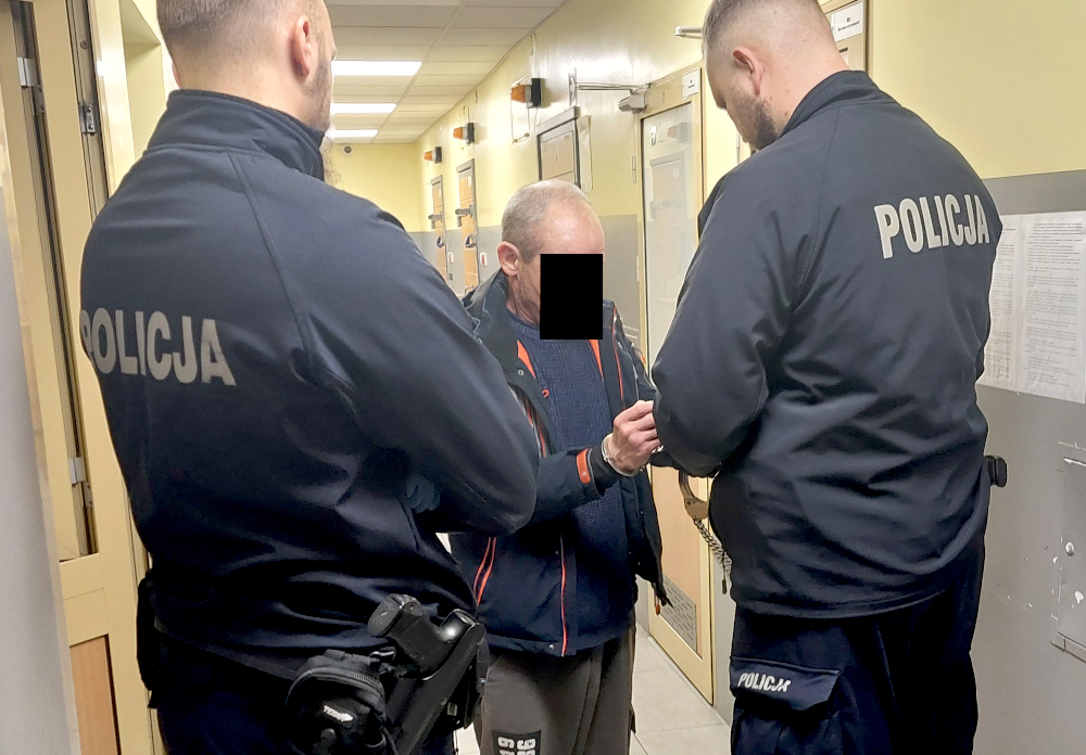 Mężczyzna w szarej bluzie stoi między dwoma policjantami w mundurach w korytarzu komisariatu.