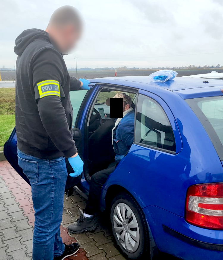 Funkcjonariusz policji w rękawiczkach kontroluje mężczyznę siedzącego w niebieskim samochodzie na parkingu.