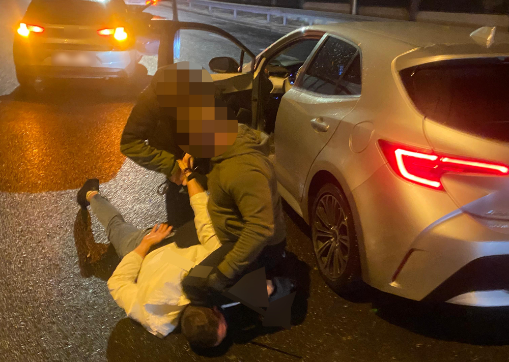Dwaj mężczyźni na jezdni trzymają jednego mężczyznę leżącego na asfalcie obok zaparkowanego samochodu.