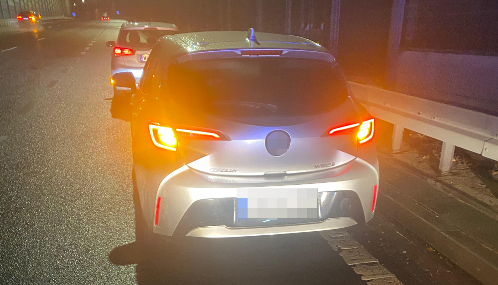 Na drodze nocą stoi srebrny samochód hybrydowy z włączonymi światłami tylnymi, za nim widoczny inny pojazd.