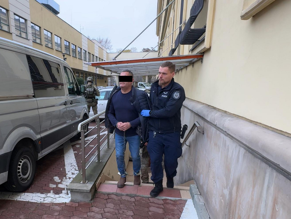 Mężczyzna w kajdankach prowadzony przez policjanta na schodach, obok stoi funkcjonariusz w mundurze.