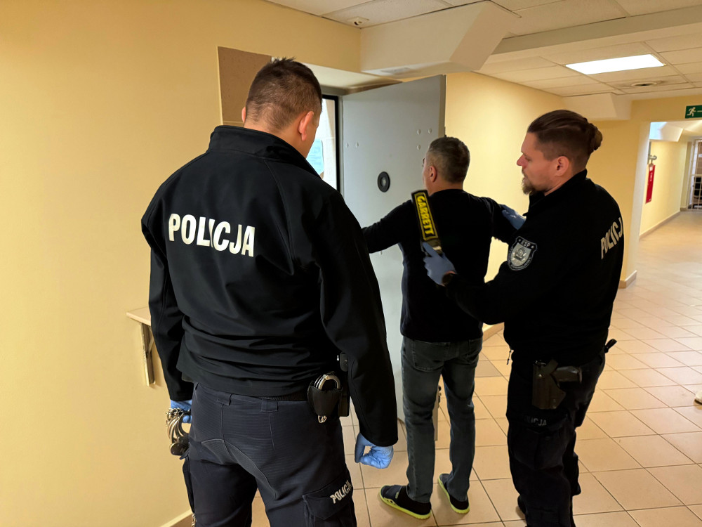 Dwóch policjantów przeszukuje mężczyznę w korytarzu budynku, używając detektora metalu.