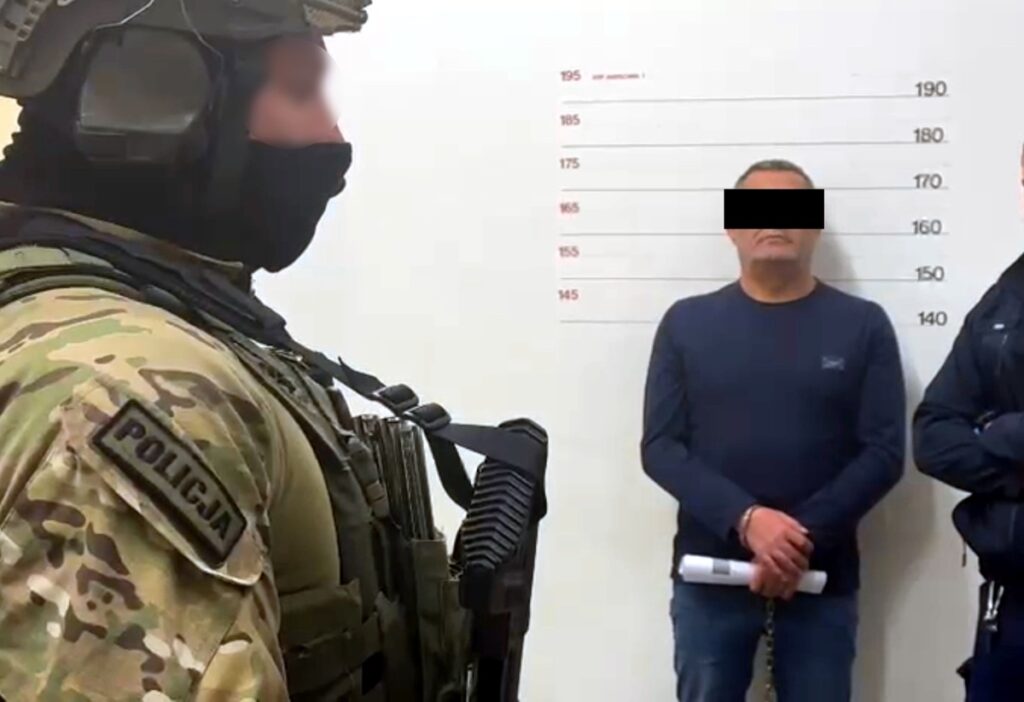 Policjant w kamizelce taktycznej stoi obok mężczyzny w kajdankach, który jest fotografowany na tle ściany z miarką.