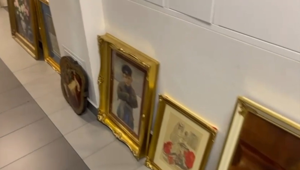 Ramy obrazów ustawione na podłodze w jasnym wnętrzu, wśród nich portret mężczyzny i ilustracja z kwiatami.