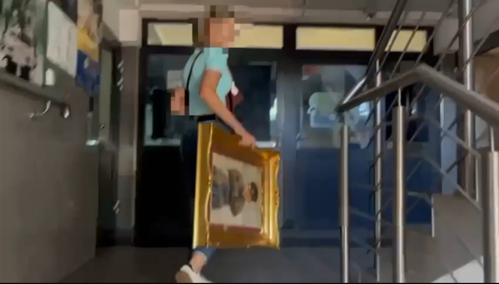 Kobieta w niebieskiej koszulce niesie w ramie obraz w holu budynku.