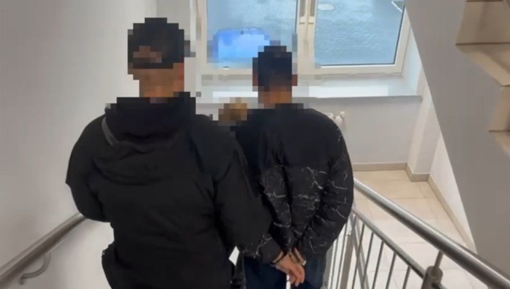 Mężczyzna w czarnej kurtce prowadzony przez funkcjonariusza policji na schodach w budynku.