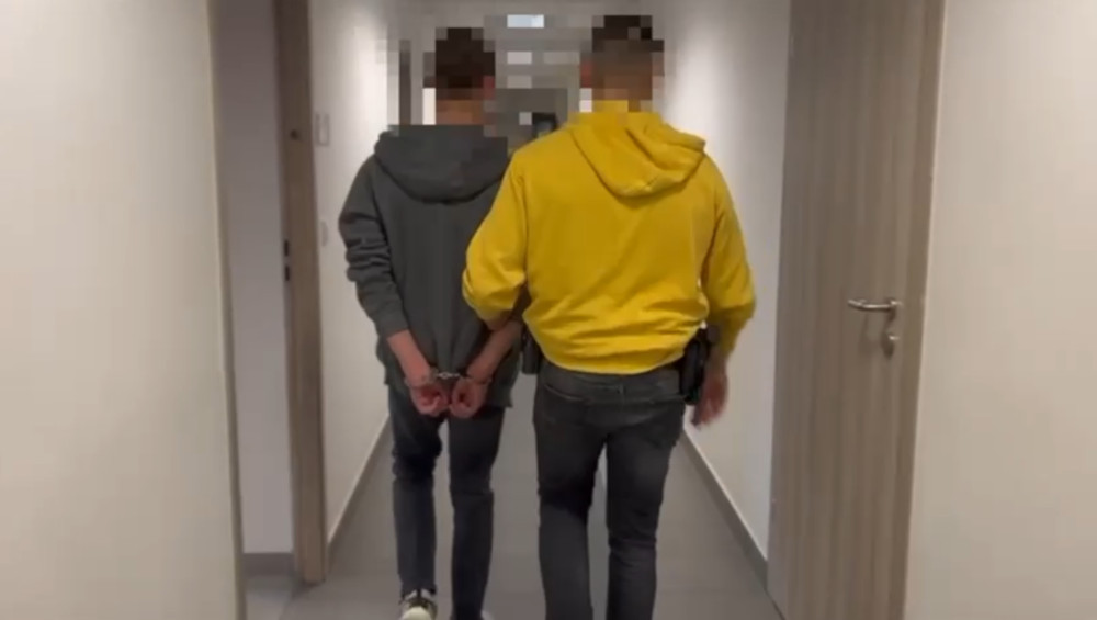 Mężczyzna w szarym dresie prowadzony przez funkcjonariusza w żółtej bluzie na korytarzu budynku.