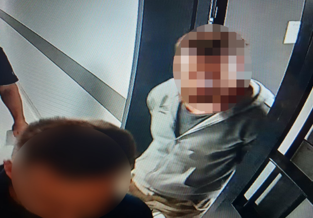 Mężczyzna w jasnej bluzie stoi w wąskim korytarzu, obok niego dwóch innych mężczyzn.