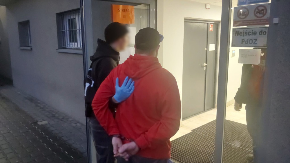 Mężczyzna w czerwonym kapturze z rękami związanymi z tyłu stoi przy drzwiach, a obok niego stoi drugi mężczyzna w czarnej bluzie.