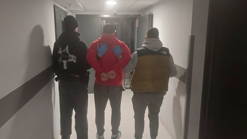 Mężczyzna w czerwonym dresie z rękami skrępowanymi z tyłu, prowadzony przez dwóch mężczyzn w korytarzu budynku.