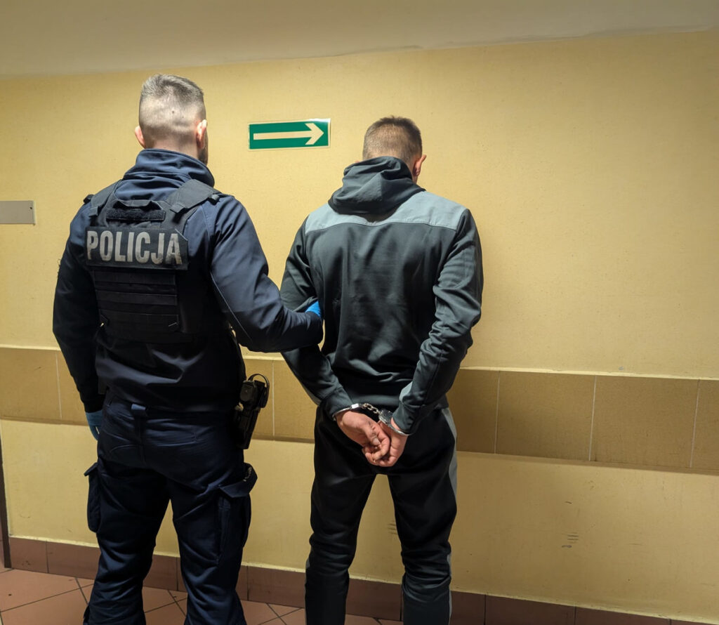 Policjant w mundurze trzyma mężczyznę w kajdankach w korytarzu z żółtymi ścianami, wskazując na wyjście.