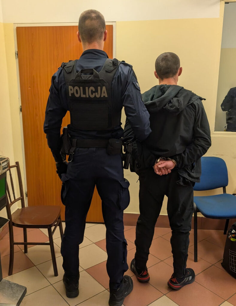 Policjant w mundurze stoi za zatrzymanym mężczyzną z rękami skrępowanymi, w pomieszczeniu z krzesłami.