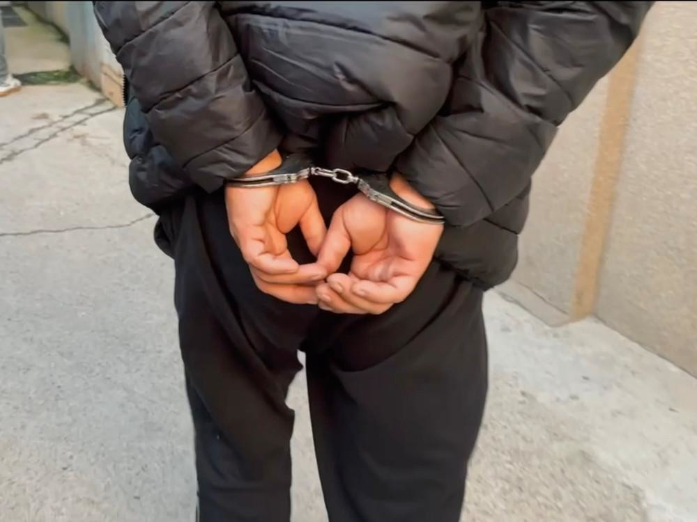 Mężczyzna w czarnej kurtce z rękami skuty w kajdankach stoi na chodniku.