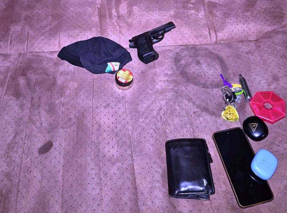 Na brązowej kanapie leży czarna broń, skarpetka, puszka, klucze, portfel oraz dwa urządzenia elektroniczne.