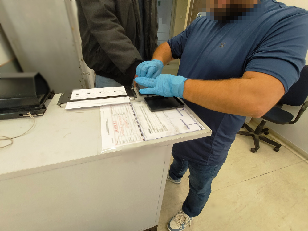 Mężczyzna w niebieskiej koszulce zakłada rękawiczki, przygotowując się do weryfikacji danych przy biurku.