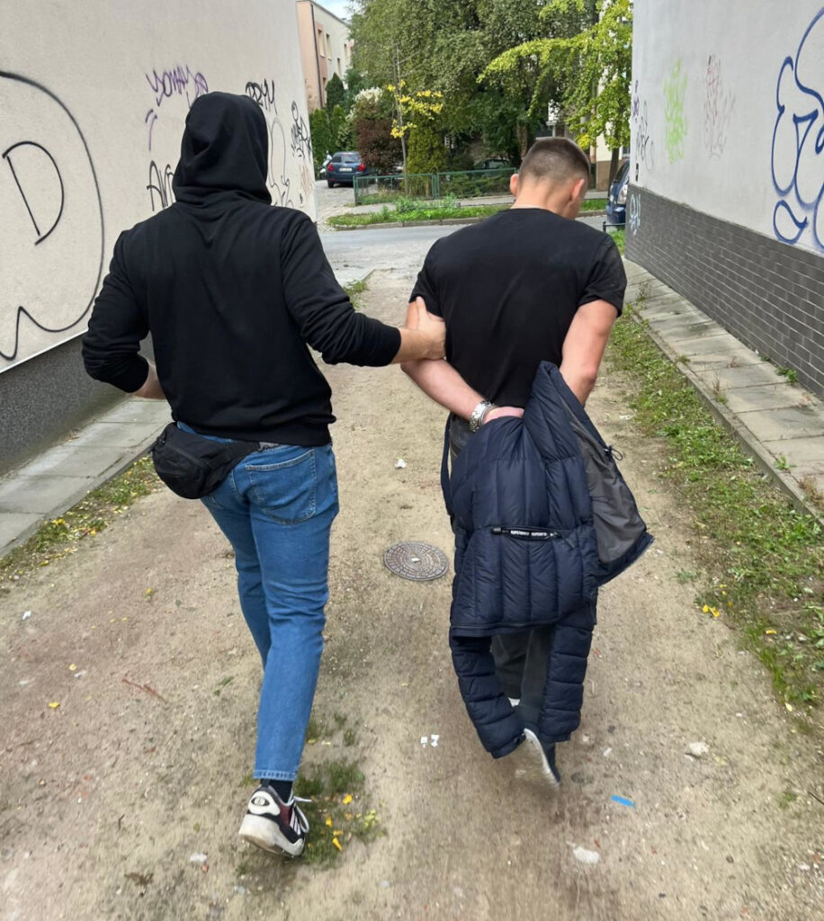 Mężczyzna w czarnej bluzie z kapturem prowadzi drugiego mężczyznę z rękami za plecami w wąskiej uliczce.