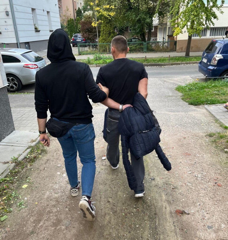 Mężczyzna w czarnej bluzie prowadzi innego mężczyznę w szarych spodniach, spacerując po chodniku w miejskim otoczeniu.