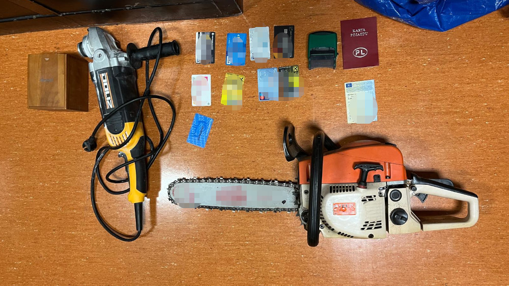 Na drewnianej powierzchni leży piła łańcuchowa, wiertarka oraz kilka kart płatniczych i dokumentów.