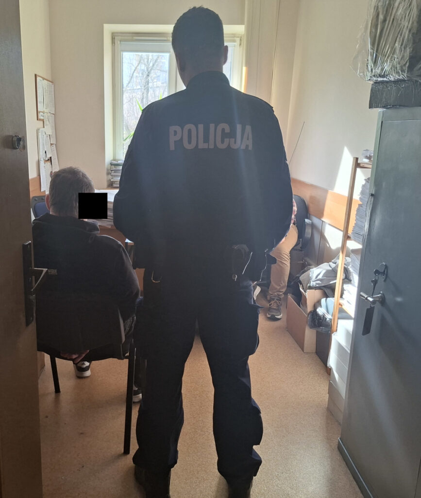 Policjant w mundurze stoi tyłem, obserwując dwóch mężczyzn siedzących w pokoju z dokumentami i meblami biurowymi.