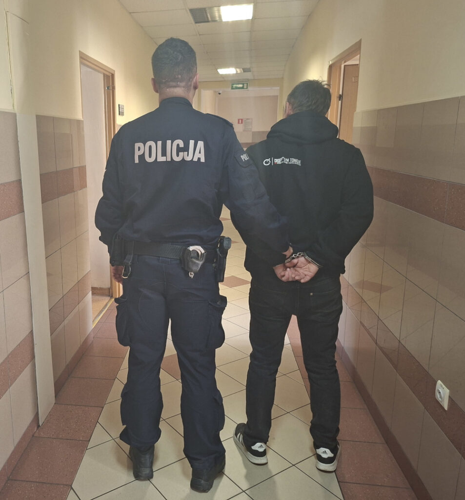 Policjant prowadzi aresztowanego mężczyznę korytarzem w budynku sądowym.