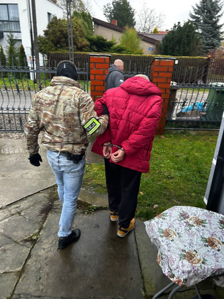 Funkcjonariusz w kamizelce taktycznej prowadzi aresztowanego mężczyznę w czerwonej kurtce przez posesję.