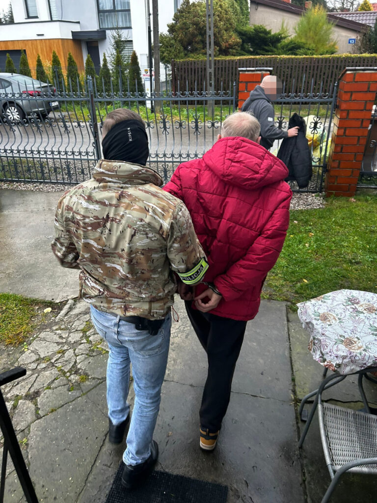 Funkcjonariusz w kamizelce policyjnej prowadzi aresztowanego mężczyznę w czerwonej kurtce przy ogrodzeniu.