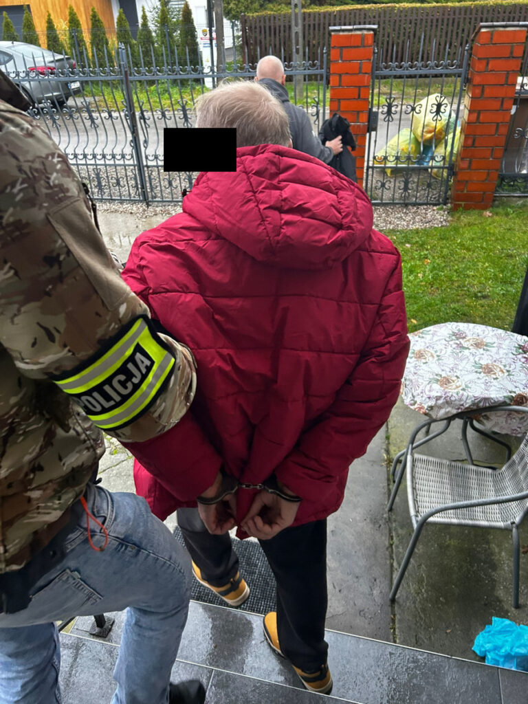 Mężczyzna w czerwonej kurtce, skuty kajdankami, prowadzony przez policjanta na schodach przed budynkiem.