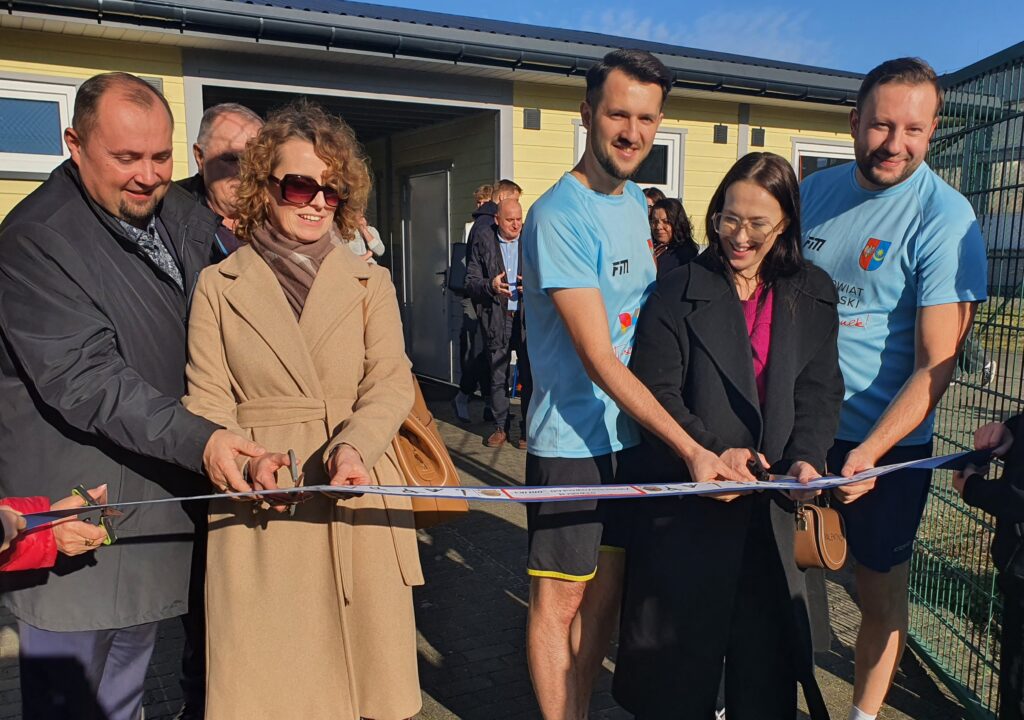 Cztery osoby trzymające nożyczki do przecięcia wstęgi na otwarciu nowego obiektu, w tle widoczna grupa gości.