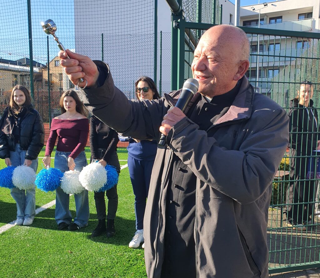 Mężczyzna z mikrofonem w ręku przemawia na boisku sportowym, otoczony przez grupę młodych kobiet z pomponami.