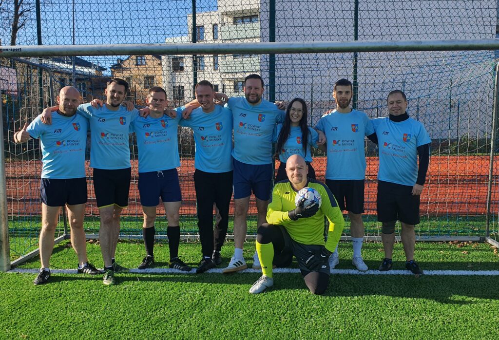 Zespół piłkarski w niebieskich koszulkach stoi przed bramką na boisku, a bramkarz w żółtym stroju klęczy z piłką.