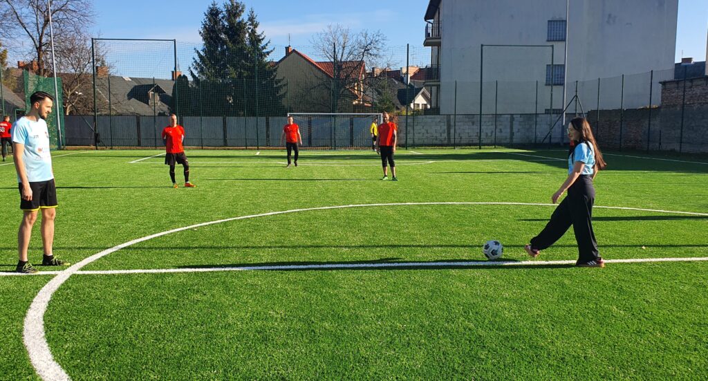 Grupa pięciu osób w czerwonych koszulkach oraz dwóch w jasnych strojach gra w piłkę nożną na boisku z sztuczną trawą.