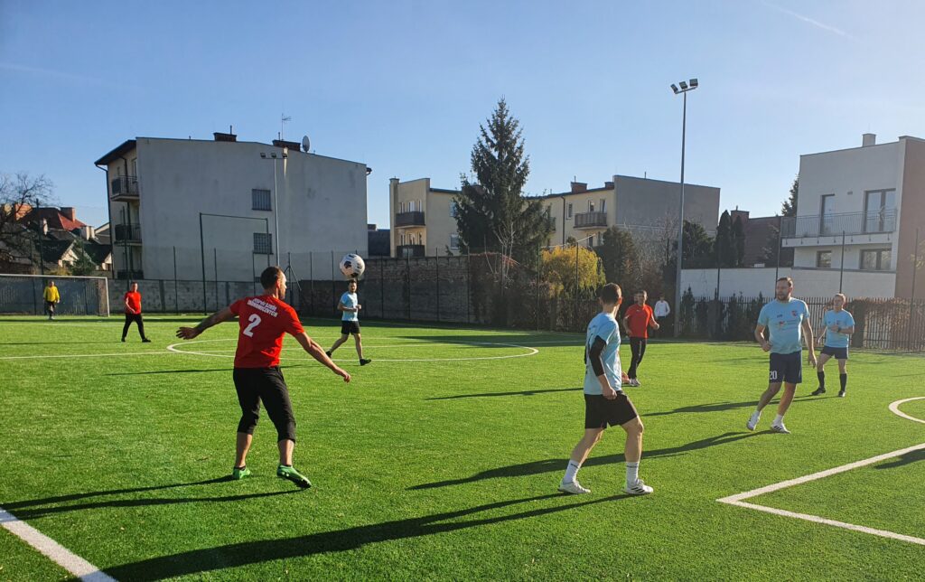 Grupa mężczyzn gra w piłkę nożną na boisku z sztuczną trawą w słoneczny dzień.