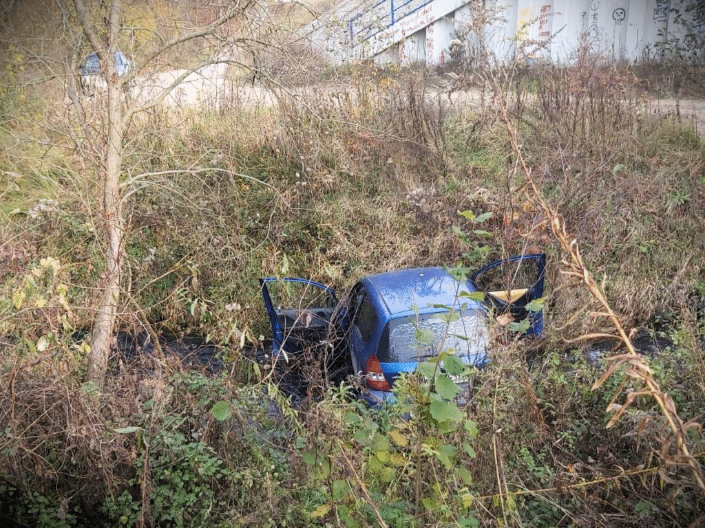 Nieoznakowany niebieski samochód wjechał do rowu, otwarte drzwi wśród wysokich traw i krzewów.