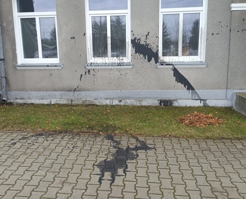 Czarna farba rozlana na ścianie budynku, z plamami spływającymi wzdłuż okien, obok trawnika z opadłymi liśćmi.