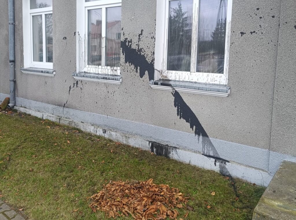 Czarna farba spływa po ścianie budynku, obok leżą uschnięte liście, a w tle widoczne są okna z zasłonami.