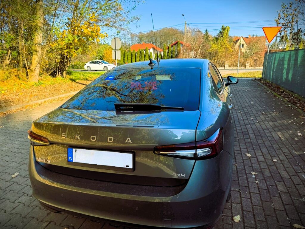 Szary samochód osobowy marki Skoda zaparkowany na brukowanej drodze w otoczeniu drzew i domów.