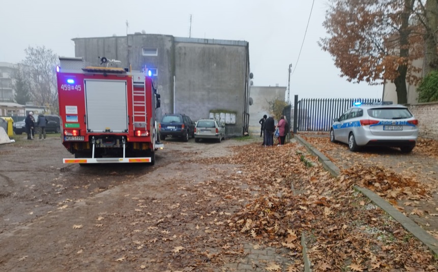 W tle szarego budynku, straż pożarna z włączonymi sygnałami świetlnymi, obok policyjny radiowóz, a w pobliżu grupka ludzi rozmawia.