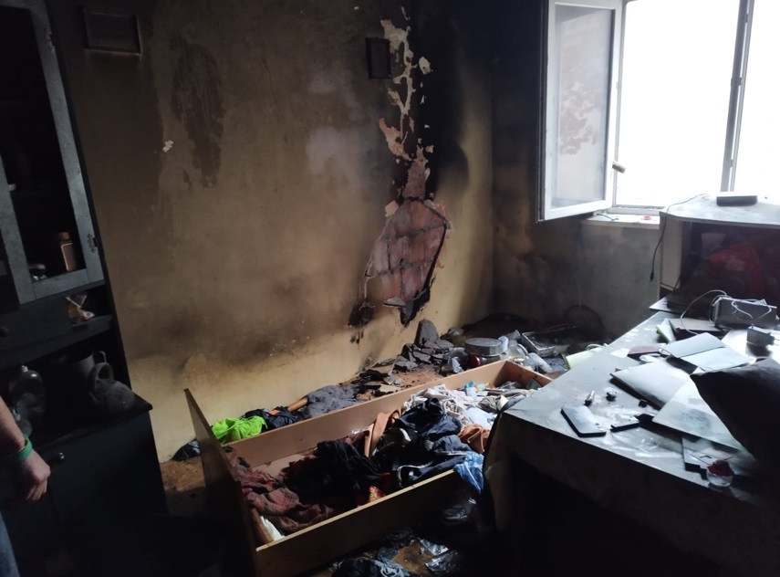 Zniszczony pokój z popalonymi ścianami, otwartym oknem i bałaganem na podłodze oraz meblach.