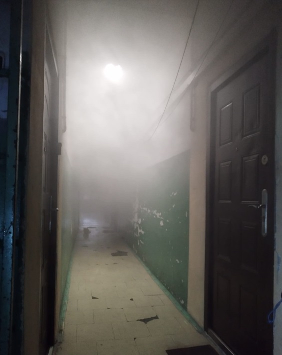 Korytarz w budynku mieszkalnym wypełniony gęstym dymem, z widocznymi drzwiami po obu stronach.