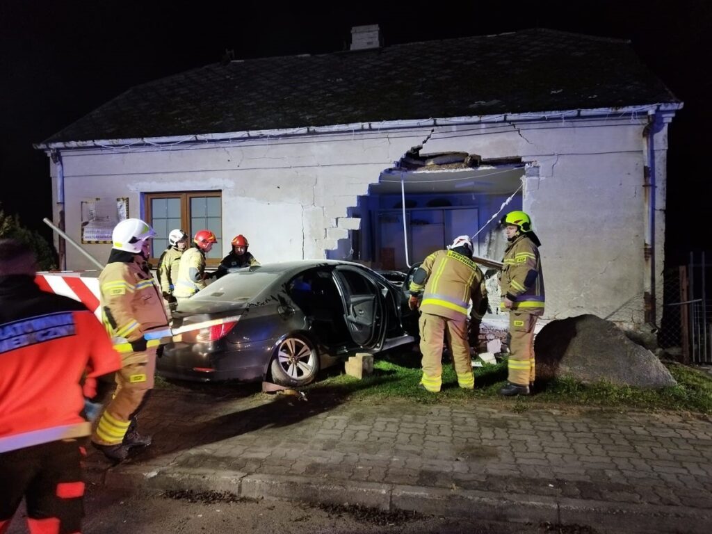 Grupa ratowników w mundurach pracuje przy uszkodzonym samochodzie, który wjechał w ścianę budynku.