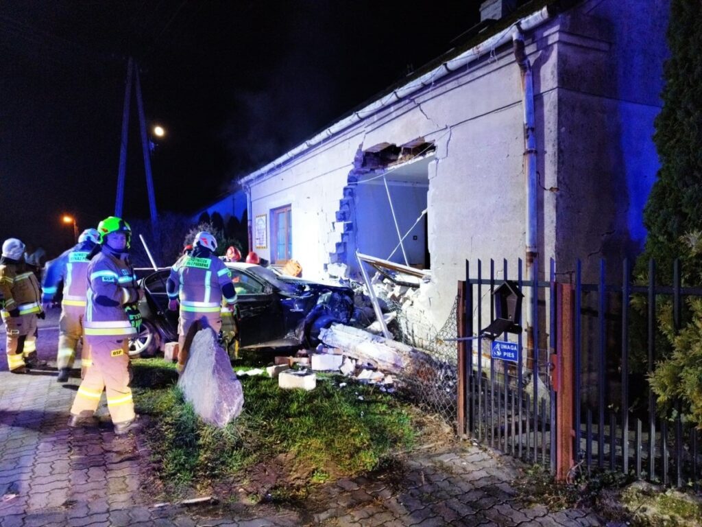 Grupa strażaków w specjalistycznym sprzęcie pracuje przy uszkodzonym budynku po wypadku samochodowym.