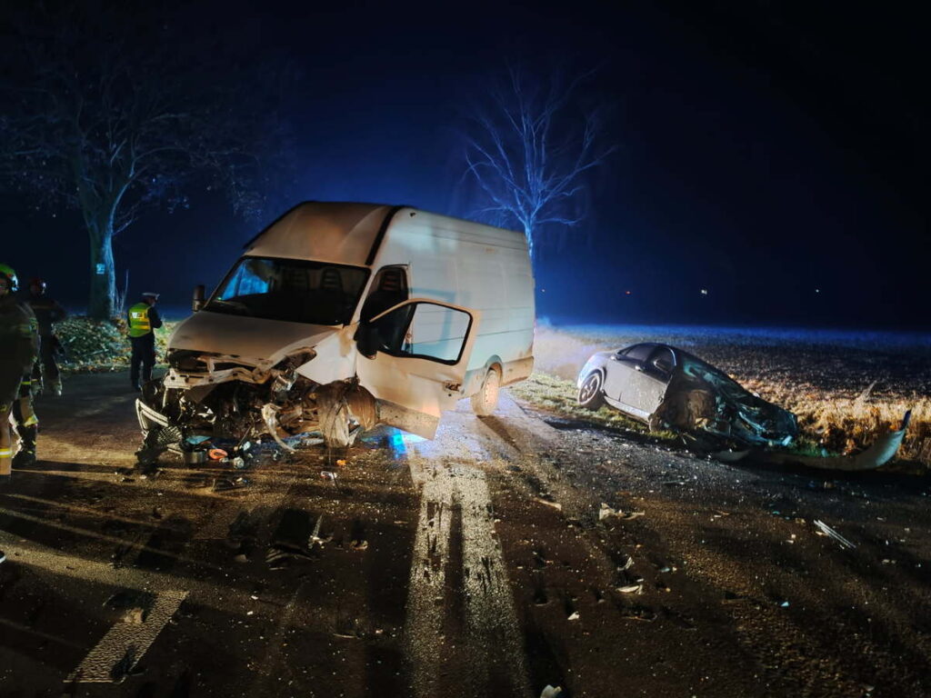 W nocy dwa rozbite pojazdy, dostawczy i osobowy, leżą na drodze, a obok pracują strażacy i policjanci.