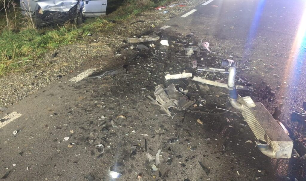 Fragment drogi pokryty szczątkami metalowymi i szkłem, obok uszkodzony samochód w tle.