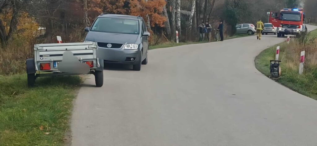 Na drodze stoi szary samochód osobowy obok przyczepy, a w tle widać straż pożarną i ludzi z rowerami.