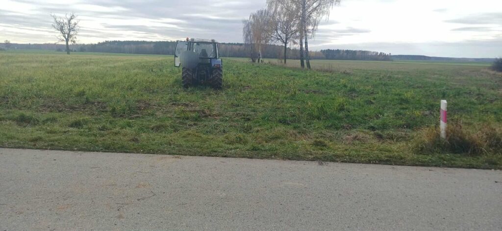Traktor pracujący na zielonym polu, w tle drzewa i chmury na niebie. Obok asfaltowa droga z oznaczeniem.